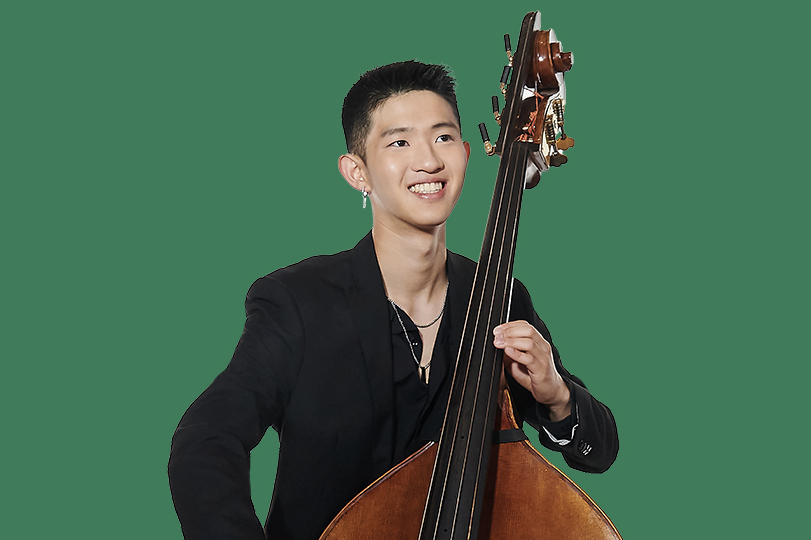 A man wearing a black shirt, holding a cello, smiling at the camera, with a dark green background.